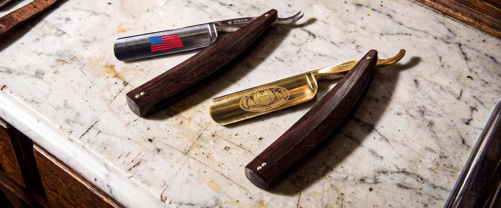The Flag Edition and the 24K Gold Amendment Edition straight razors of the Declaration of Independence Collection on a marble surface